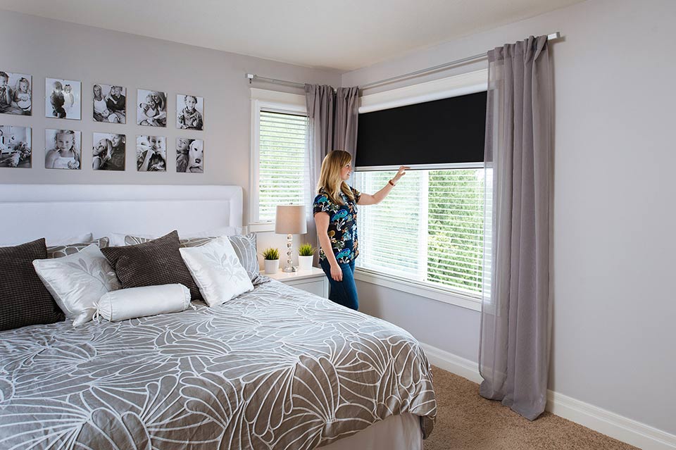 Woman lowering a retractable window shade in a cozy, light-filled bedroom for privacy and light control.