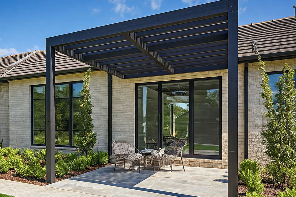 Modern patio with black metal pergola structure attached to light brick house, featuring outdoor seating and landscaped garden.