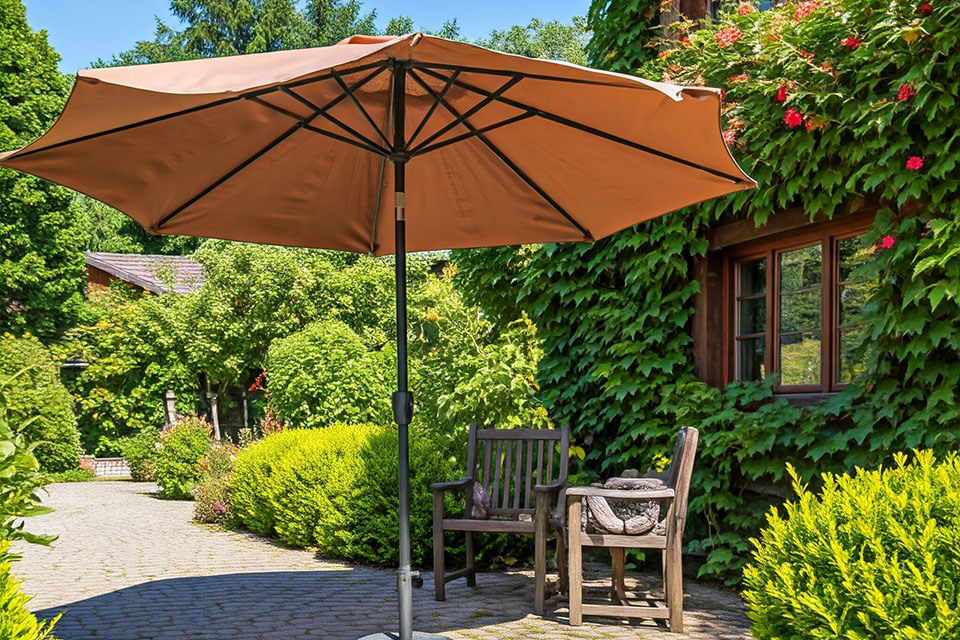 Garden patio with brown umbrella shading wooden chairs, surrounded by ivy-covered walls and colorful flowering plants.