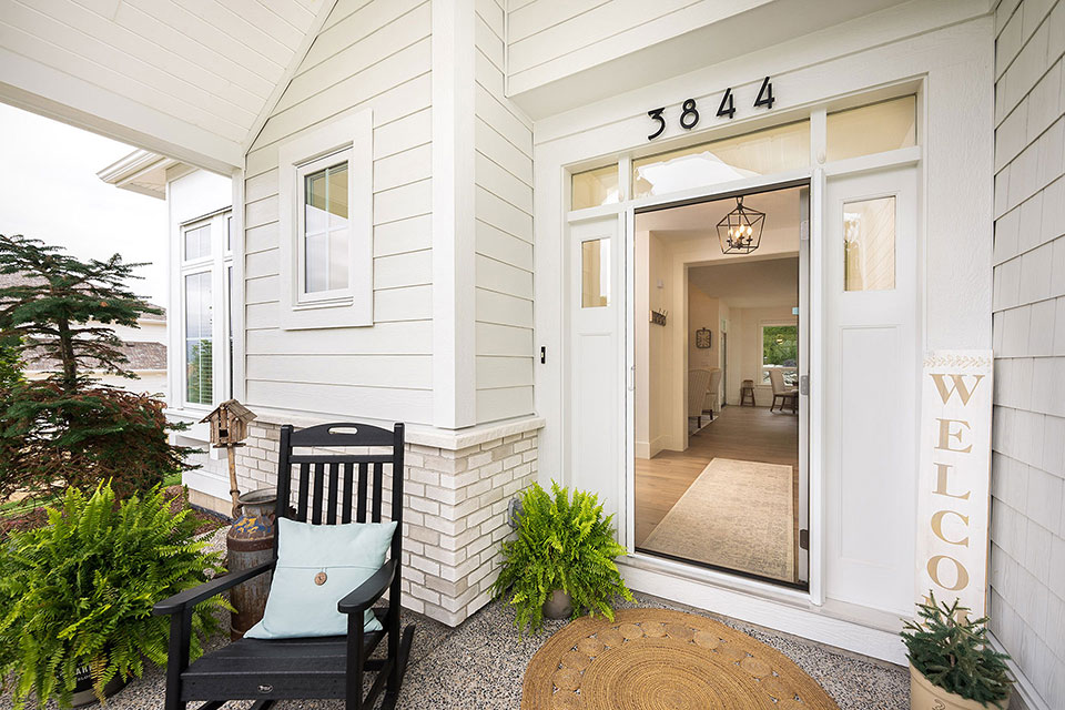 Modern white home entrance with retractable screen door, rocking chair, and potted ferns, showing lantern light inside.