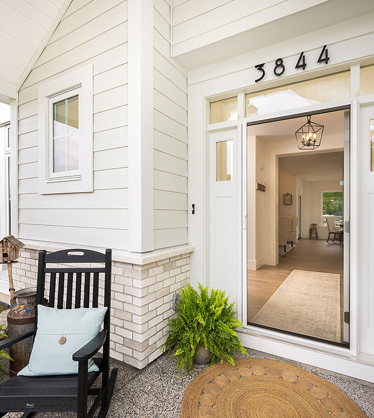 Modern white home entrance with retractable screen door, rocking chair, and potted ferns, showing lantern light inside.